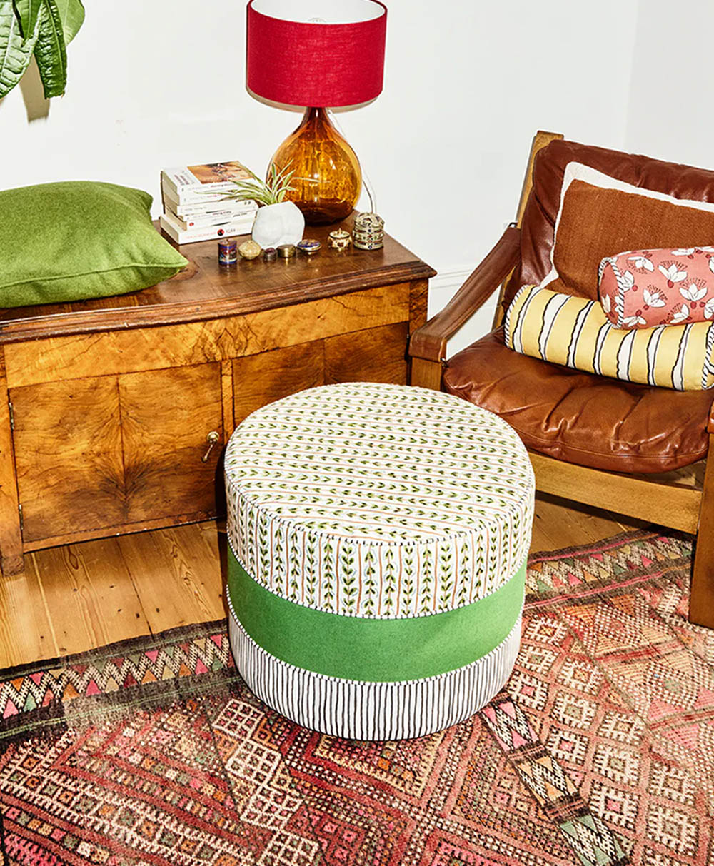 A cozy living room with a brown wooden armchair, patterned cushions, a round ottoman with green and striped sections, a colorful rug, a wooden chest, books, and an amber lamp with a red shade.