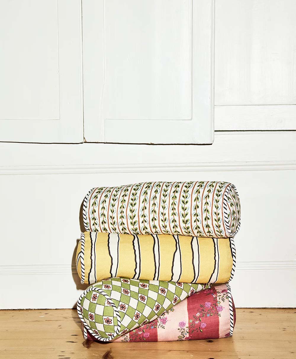Three patterned quilts are rolled and stacked on a wooden floor against a white wall. The top quilt is white with green vines, the middle is yellow with black wavy lines, and the bottom has a green and red geometric design.