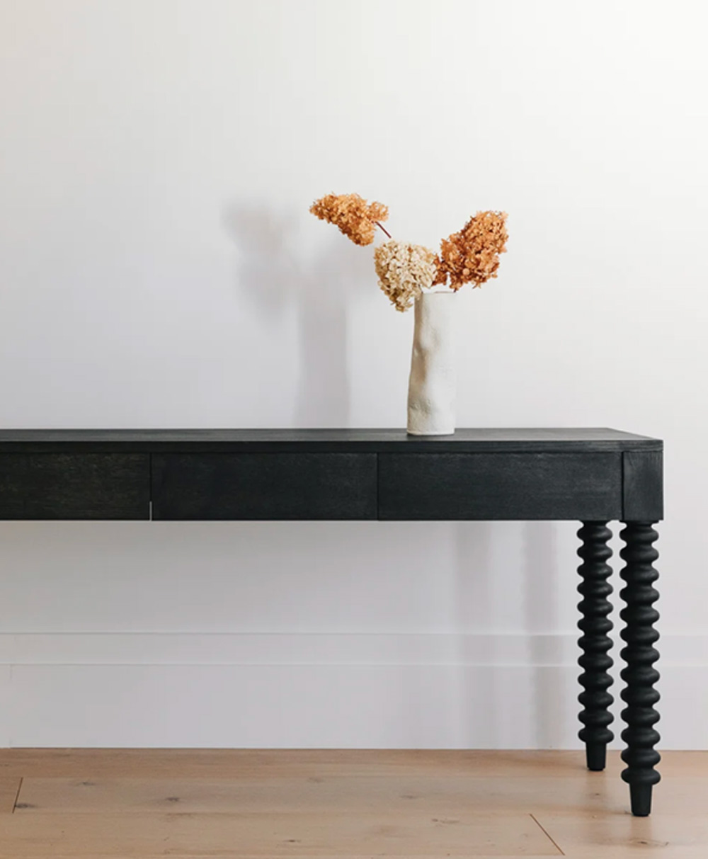 A black console table with spiral legs holds a white vase containing dried orange and beige flowers, set against a plain white wall and light wooden floor.