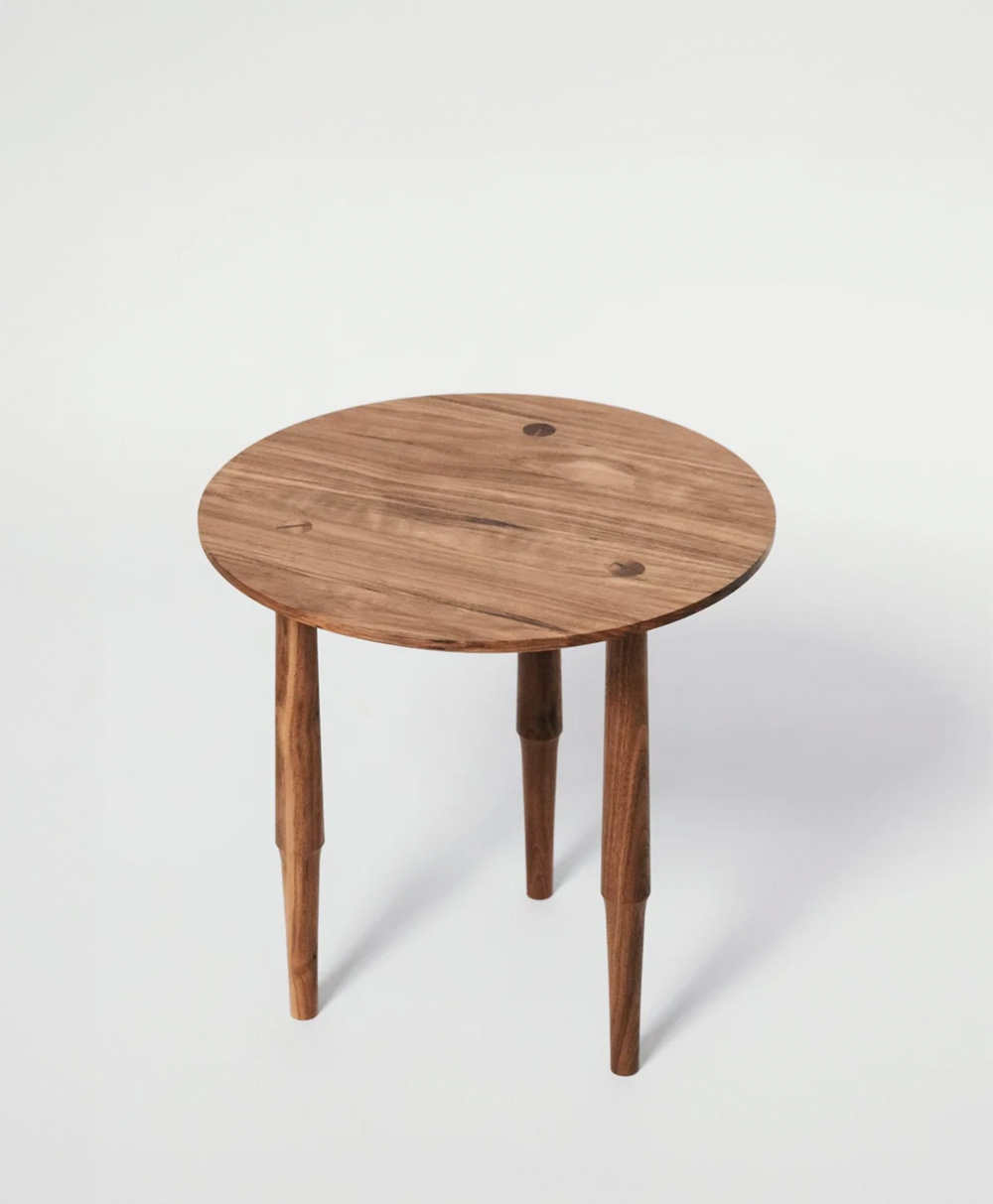 A small, round wooden table with three tapered legs, set against a plain white background. The table has a natural wood grain finish and a minimalist design.