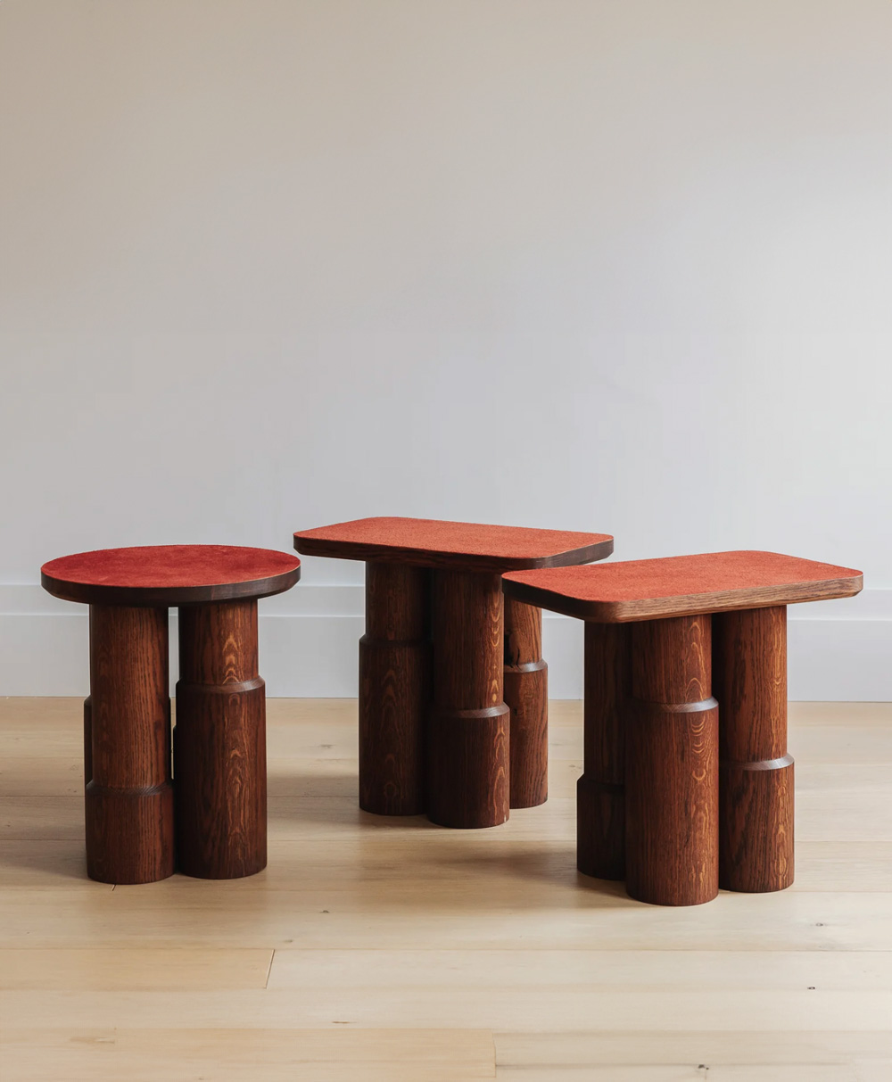 Three modern wooden tables with reddish-brown tops and cylindrical bases are arranged on a light wood floor against a plain light-colored wall.