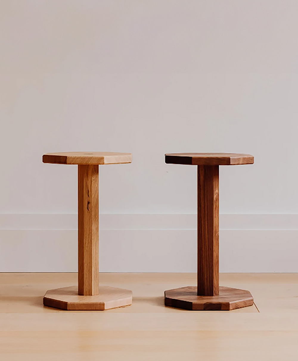 Two wooden stools with hexagonal tops and bases stand side by side on a light wood floor against a plain, light-colored wall. One stool is light wood, the other is darker wood.