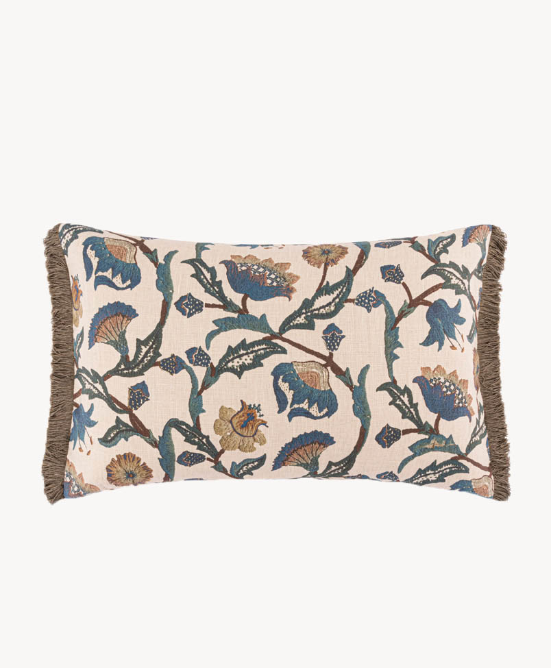 A rectangular cushion with a beige background, decorated with a botanical and floral pattern in blue, green, and brown tones, and finished with brown fringe trim along the edges.