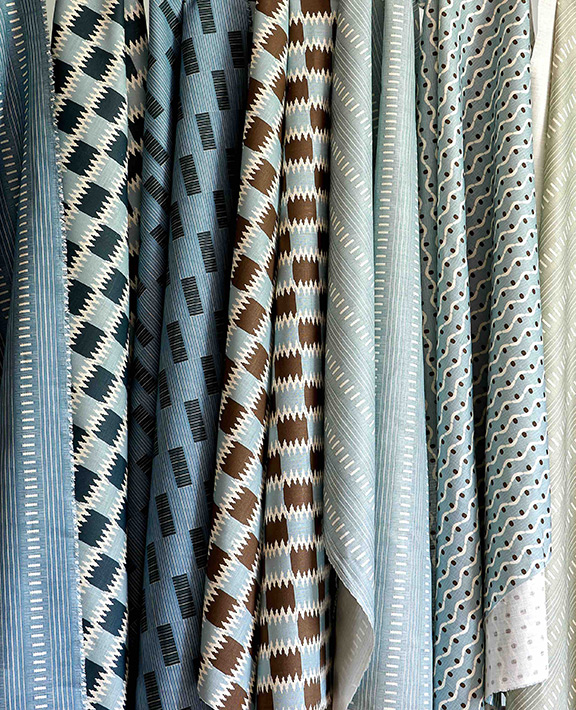 Several pieces of fabric in blue, brown, and white tones hang vertically, each featuring geometric patterns such as stripes, diamonds, and zigzags. The designs vary but share a cohesive color palette.