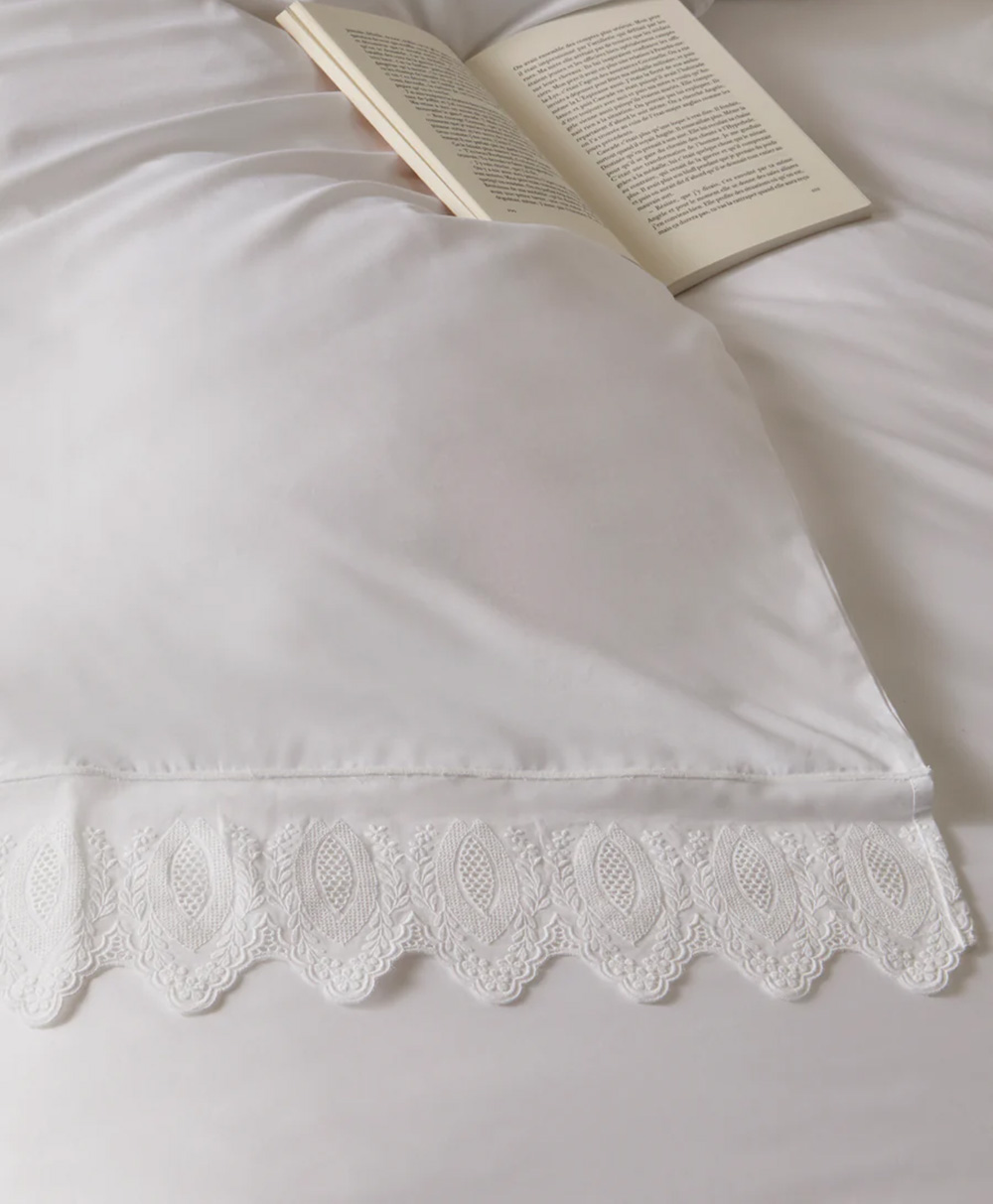 A white pillow with decorative lace trim rests on a neatly made bed, with an open book placed nearby on the white bedding.