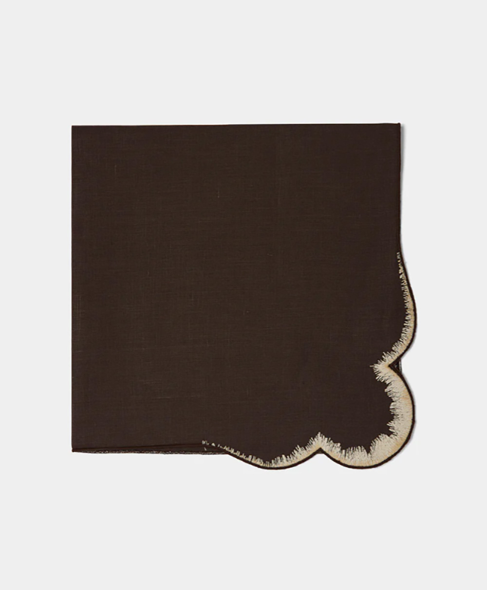 A dark brown square napkin with one scalloped corner outlined in beige embroidery, displayed flat against a white background.