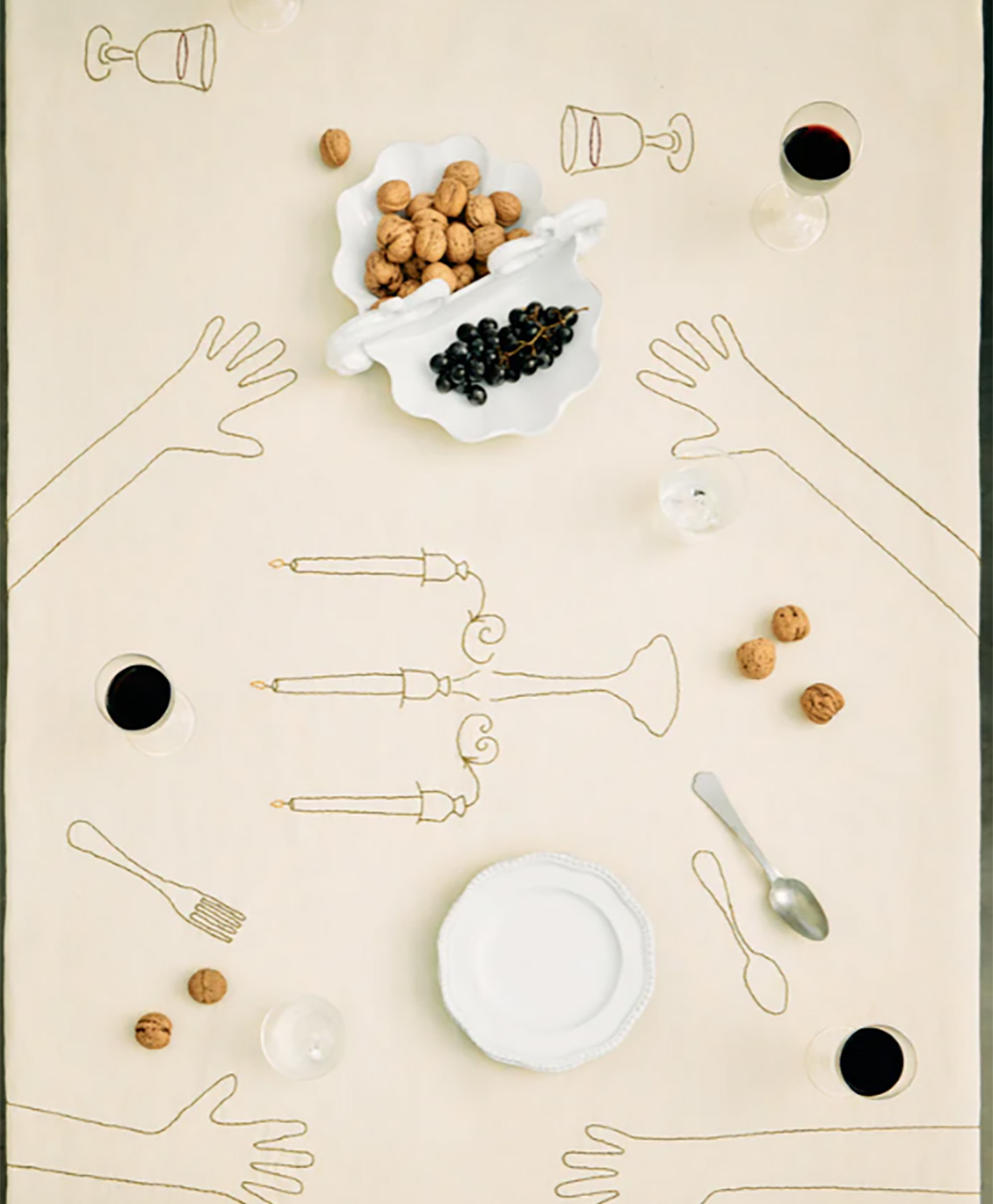 A table set with a cream tablecloth featuring embroidered outlines of hands, candles, and utensils. There are wine glasses, a dish with walnuts and grapes, scattered walnuts, and empty plates and cutlery.