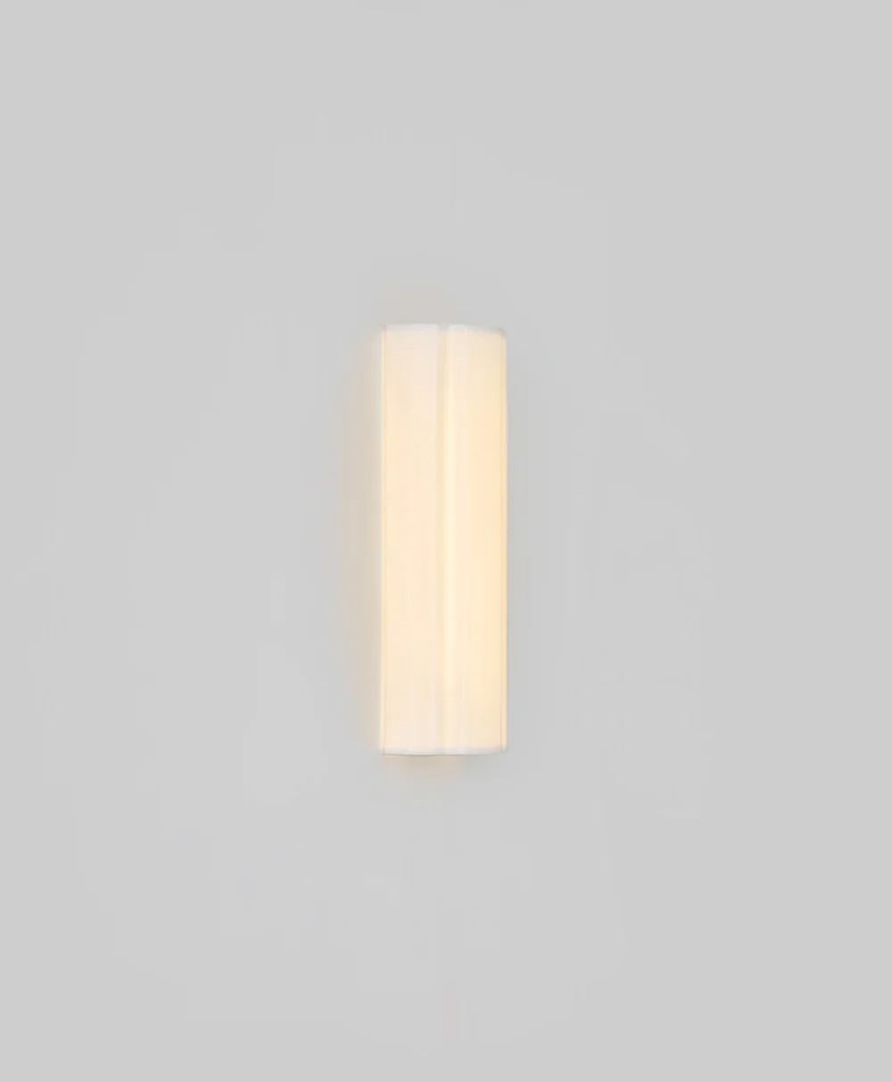 A minimalist, cylindrical wall sconce with a soft, warm glow is mounted on a plain light gray wall. The fixture has a smooth, white surface and emits light evenly, creating a modern and clean look.