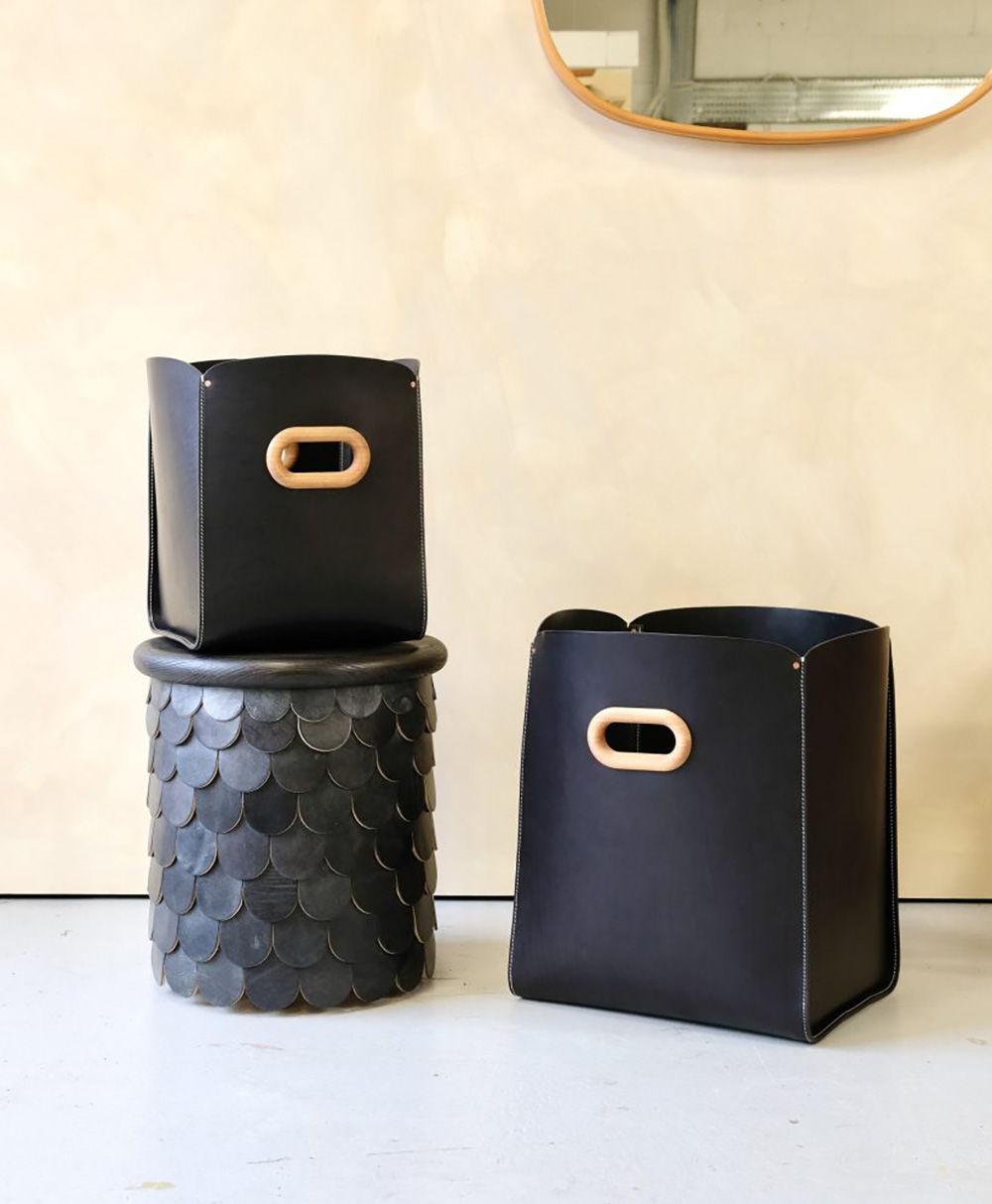 Two black leather storage bins with wooden handles are displayed; one sits atop a round, textured black stool with a scale pattern, and the other is placed on the floor, in front of a beige wall and a partial mirror.