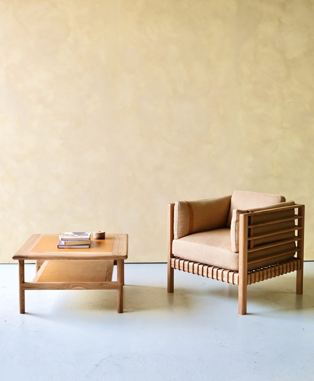 A minimalist room features a wooden armchair with beige cushions next to a matching wooden coffee table, which holds a stack of books and a small cup. The background is a light beige wall.