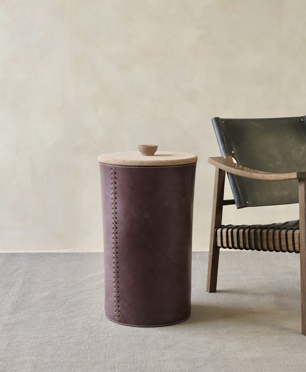 A round, brown leather side table with a wooden lid stands beside a modern wooden chair with a black leather seat and woven details, set against a neutral wall and light carpeted floor.