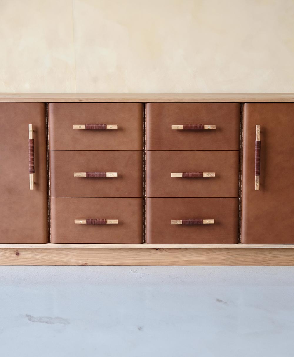 A brown leather cabinet with six central drawers and two side doors, each featuring wooden handles wrapped with a darker material, standing on a light wood base against a beige wall.
