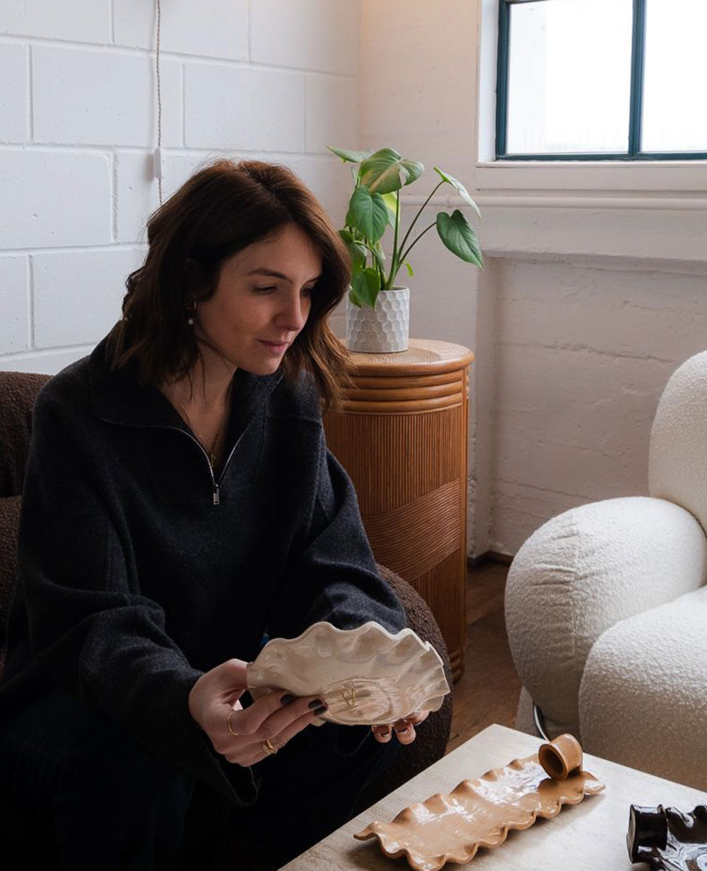A woman with brown hair sits on a dark sofa in a cozy room, holding a wavy ceramic dish and looking at another on a small table. A green potted plant sits on a wooden cabinet behind her near a window.