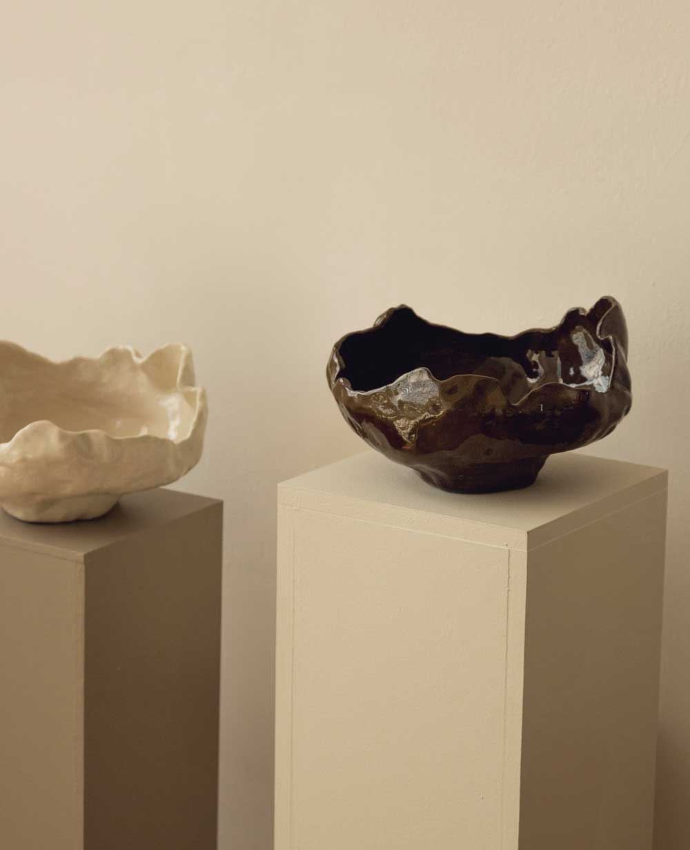 Two ceramic bowls with irregular, organic shapes are displayed on separate beige pedestals. The bowl on the left is light-colored, while the one on the right is dark and glossy. The background is neutral.