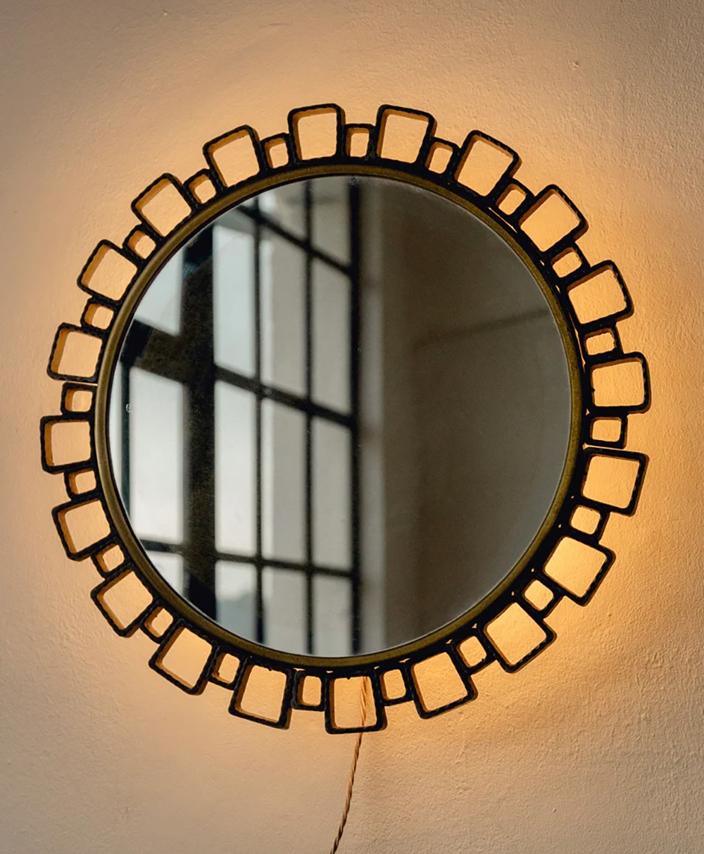 A round mirror with a decorative geometric frame hangs on a light-colored wall, reflecting a window with multiple panes. A warm light glows around the mirror’s edges.