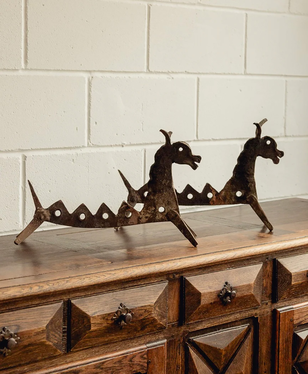 Two metal objects shaped like stylized dragons with holes along their bodies sit on a carved wooden dresser against a white brick wall.
