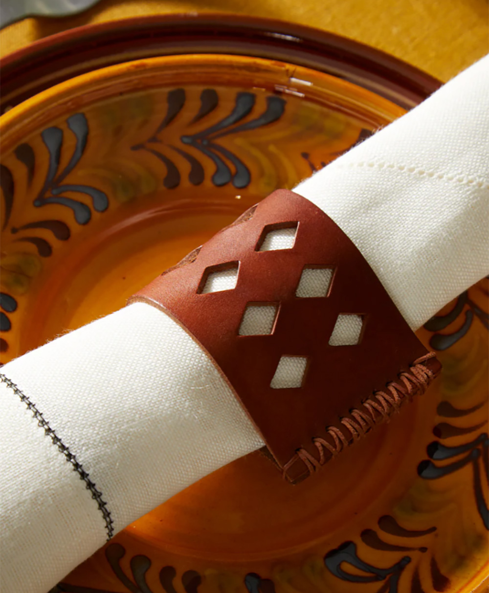 A white cloth napkin is held by a brown leather napkin ring with diamond-shaped cutouts, resting on an orange ceramic plate with blue and black decorative patterns.