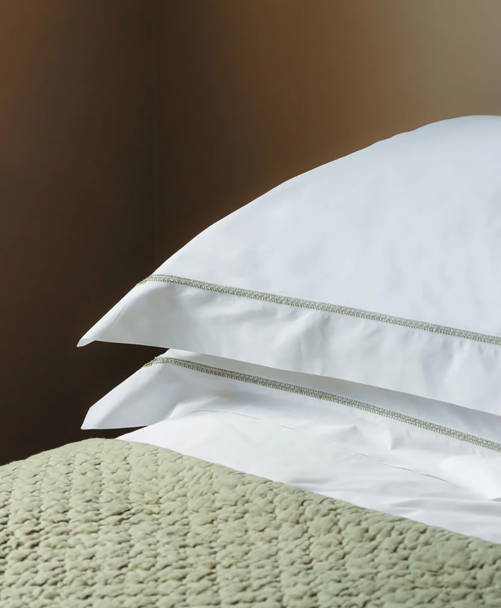 Two stacked white pillows with subtle decorative edging rest on a neatly made bed with a textured, pale green blanket, set against a warm brown wall.