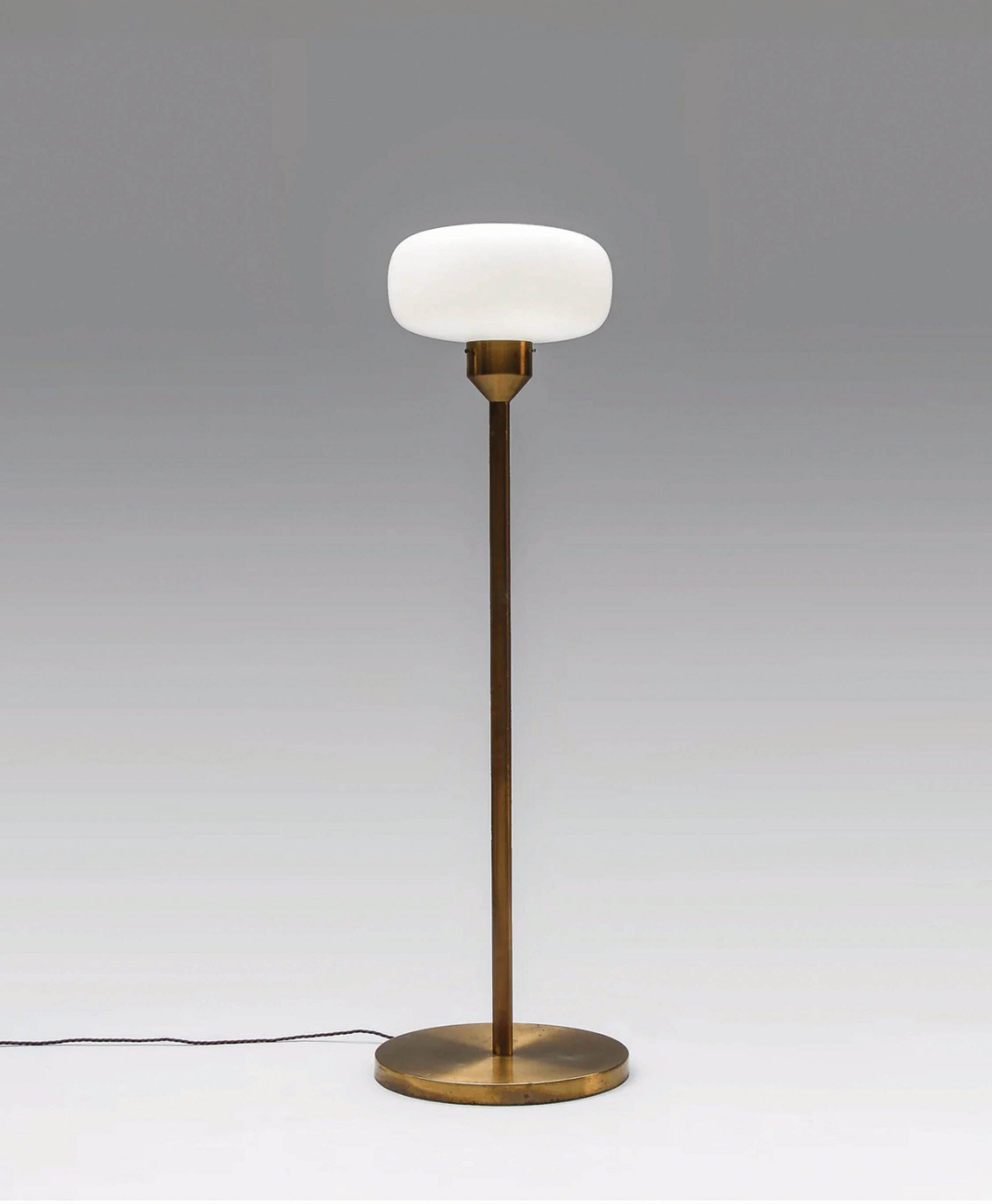 A modern floor lamp with a round white glass shade and a slender brass stand, set on a circular brass base with a visible power cord on a plain grey background.