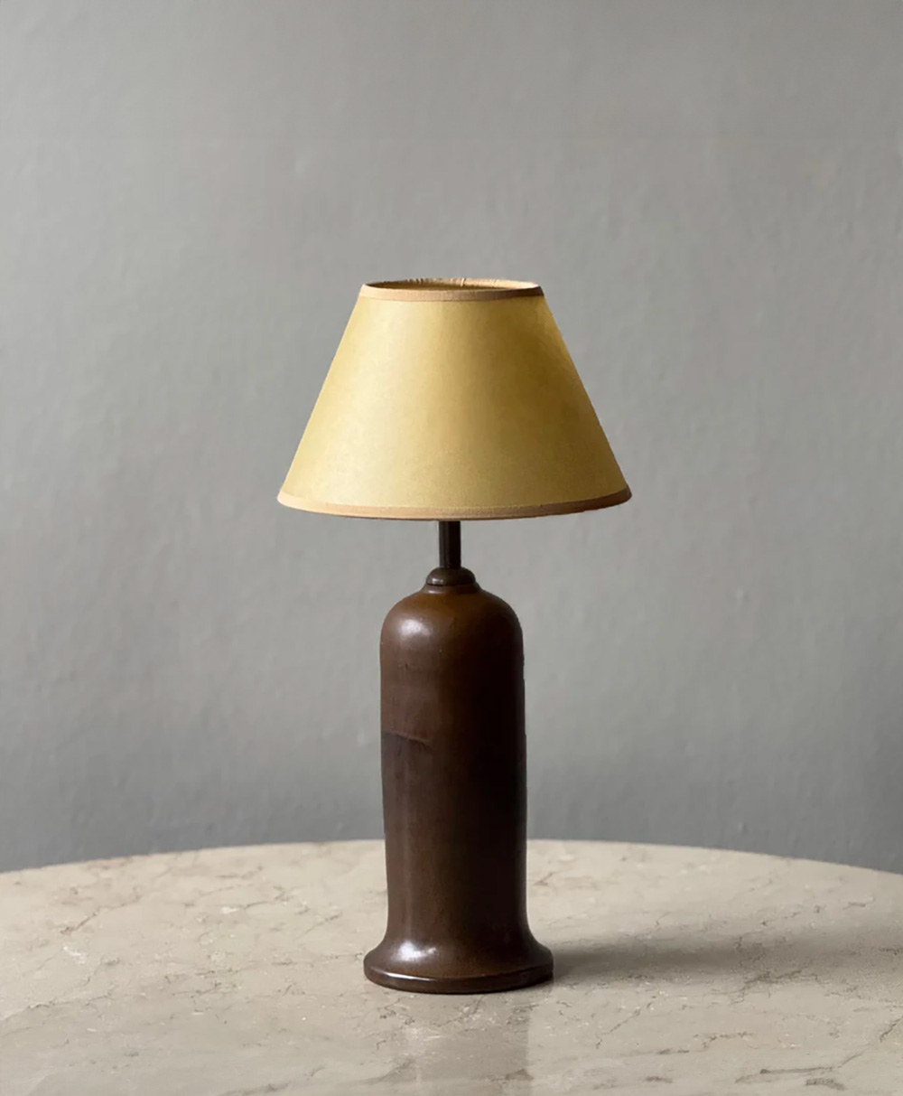 A brown table lamp with a beige lampshade sits on a light marble surface against a plain gray background.