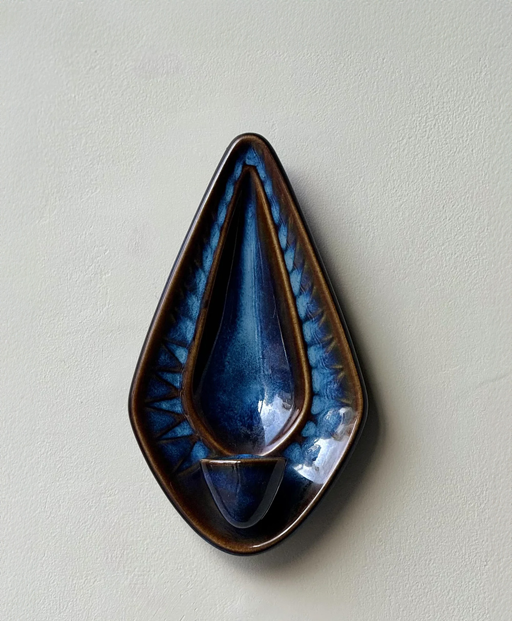 A ceramic wall sculpture with a teardrop shape, featuring a glossy blue interior and a brown rim. The design includes textured, angled patterns radiating from the center. It is displayed on a plain, light-colored wall.