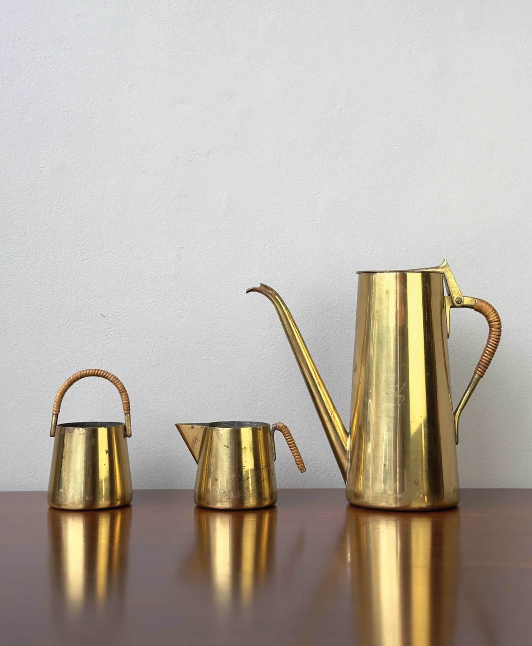Three gold-toned metal vessels—a small teapot, a creamer, and a tall coffee pot with a long spout—sit on a wooden surface against a plain light gray wall. The handles are wrapped in light-colored material.
