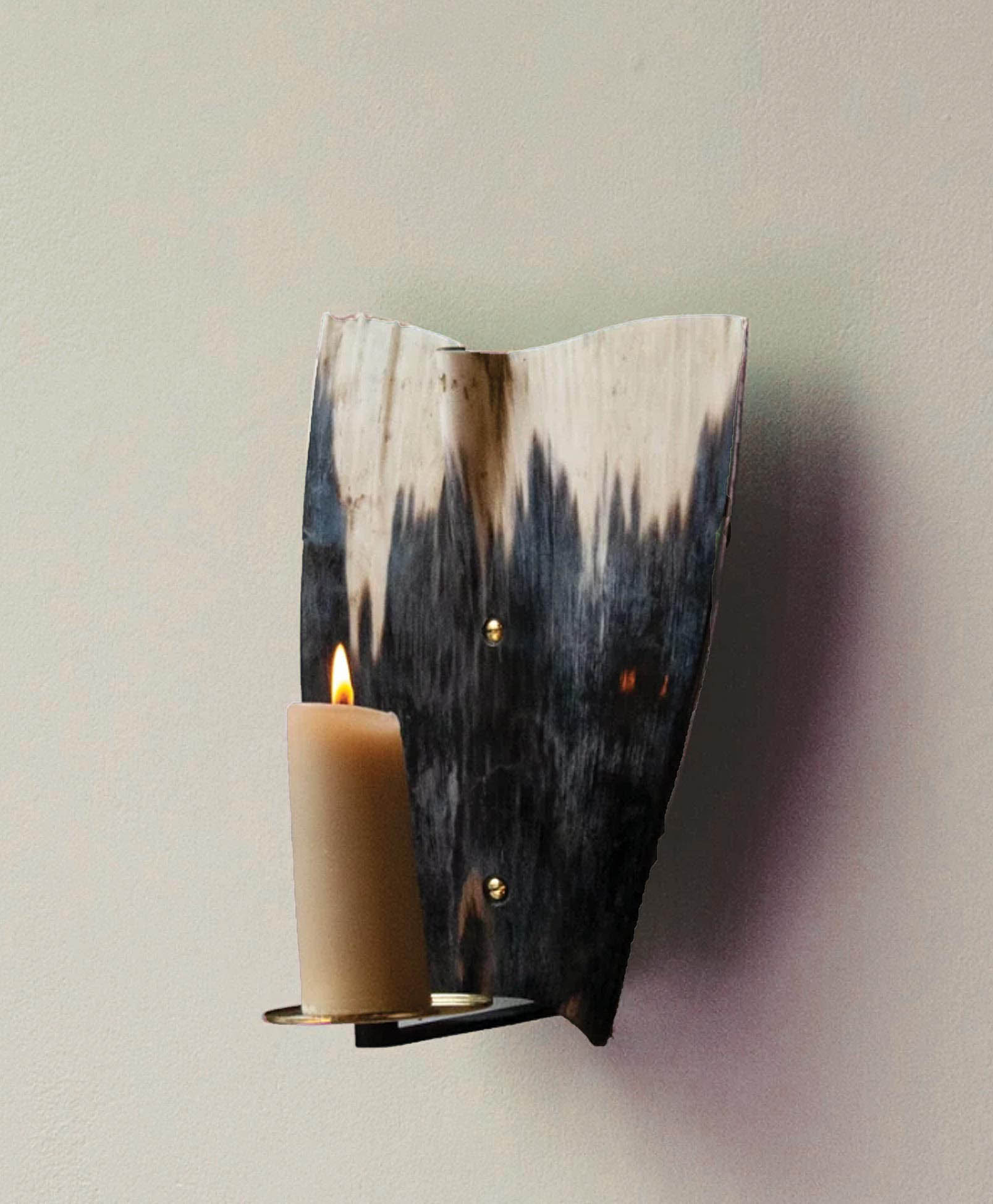 A lit candle sits on a metal holder attached to a wall-mounted sconce made from a curved, polished animal horn with dark and light patterns.