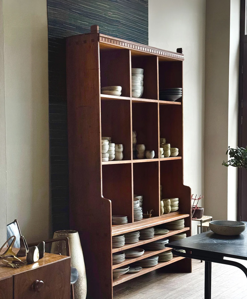 A tall wooden shelving unit displays neatly stacked ceramic dishes, bowls, and cups in a softly lit room with neutral walls and minimalist decor.