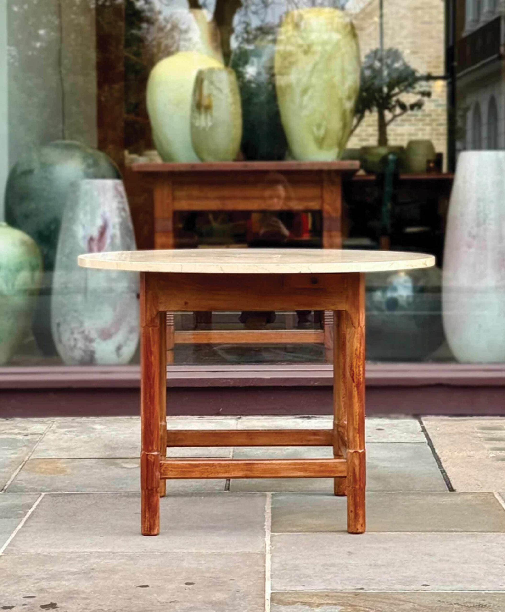 A round wooden table stands on a stone patio in front of a large window displaying oversized ceramic vases and jars inside.