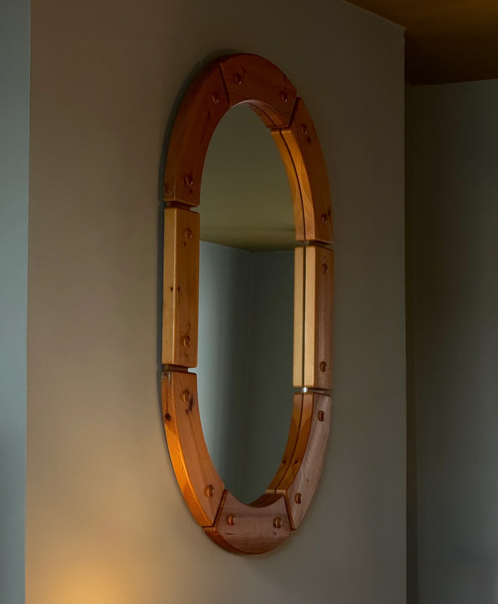 A wooden-framed oval mirror hangs on a gray wall, reflecting part of an adjacent room. Warm light softly illuminates the frame, highlighting the natural wood grain and knots.