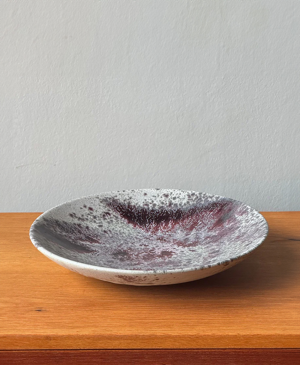 A ceramic plate with a speckled gray and deep maroon pattern sits on a wooden surface against a plain light gray wall.