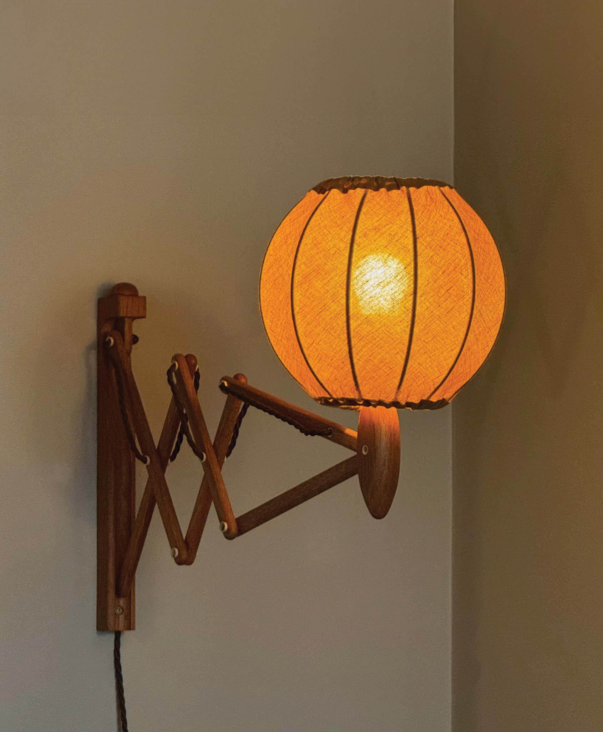 A wooden wall-mounted lamp with an accordion-style adjustable arm and a round, orange fabric lampshade is lit, casting a warm glow against a neutral wall.