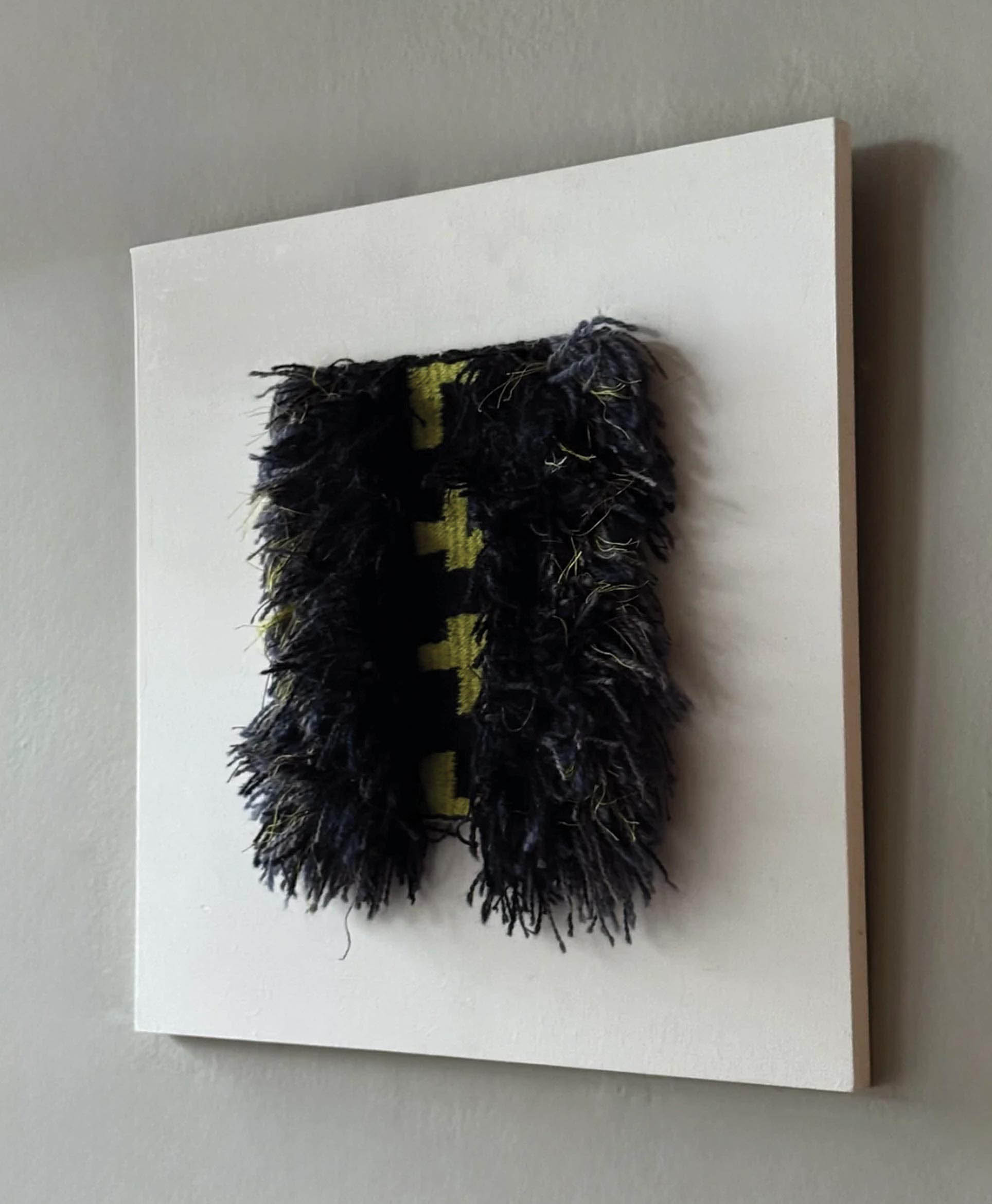 A textured art piece with shaggy, dark blue and black fibers hangs on a plain white square board. Yellow geometric shapes are partially visible beneath the fibers. The board is mounted on a gray wall.