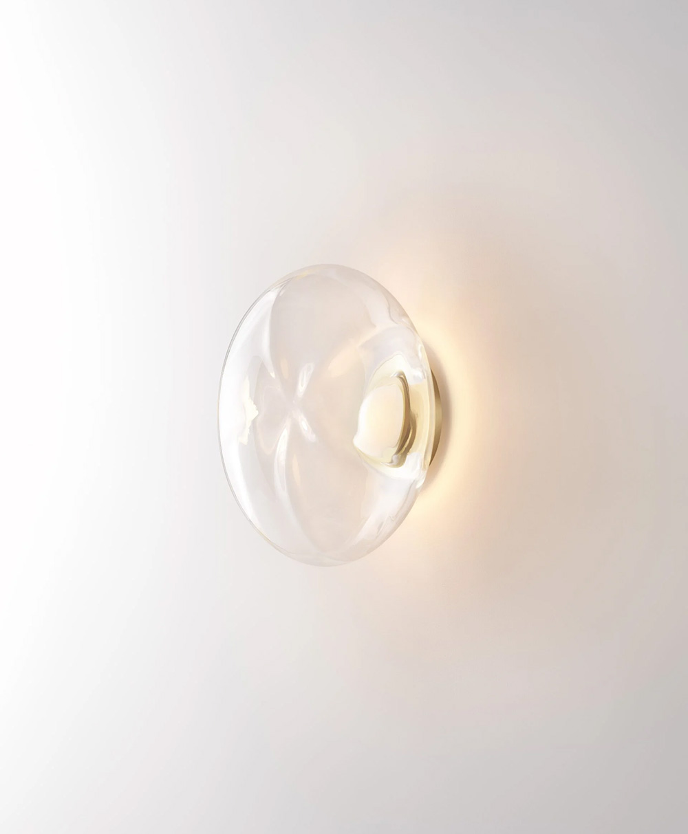 A round, clear glass wall sconce with a soft warm light glows against a plain white wall. The fixture appears minimalistic and modern.