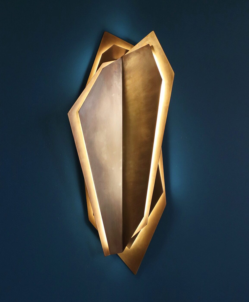 A modern wall sconce with angular, geometric metallic panels emits a warm light, casting soft glows on a dark blue wall. The design resembles overlapping shards or crystals.