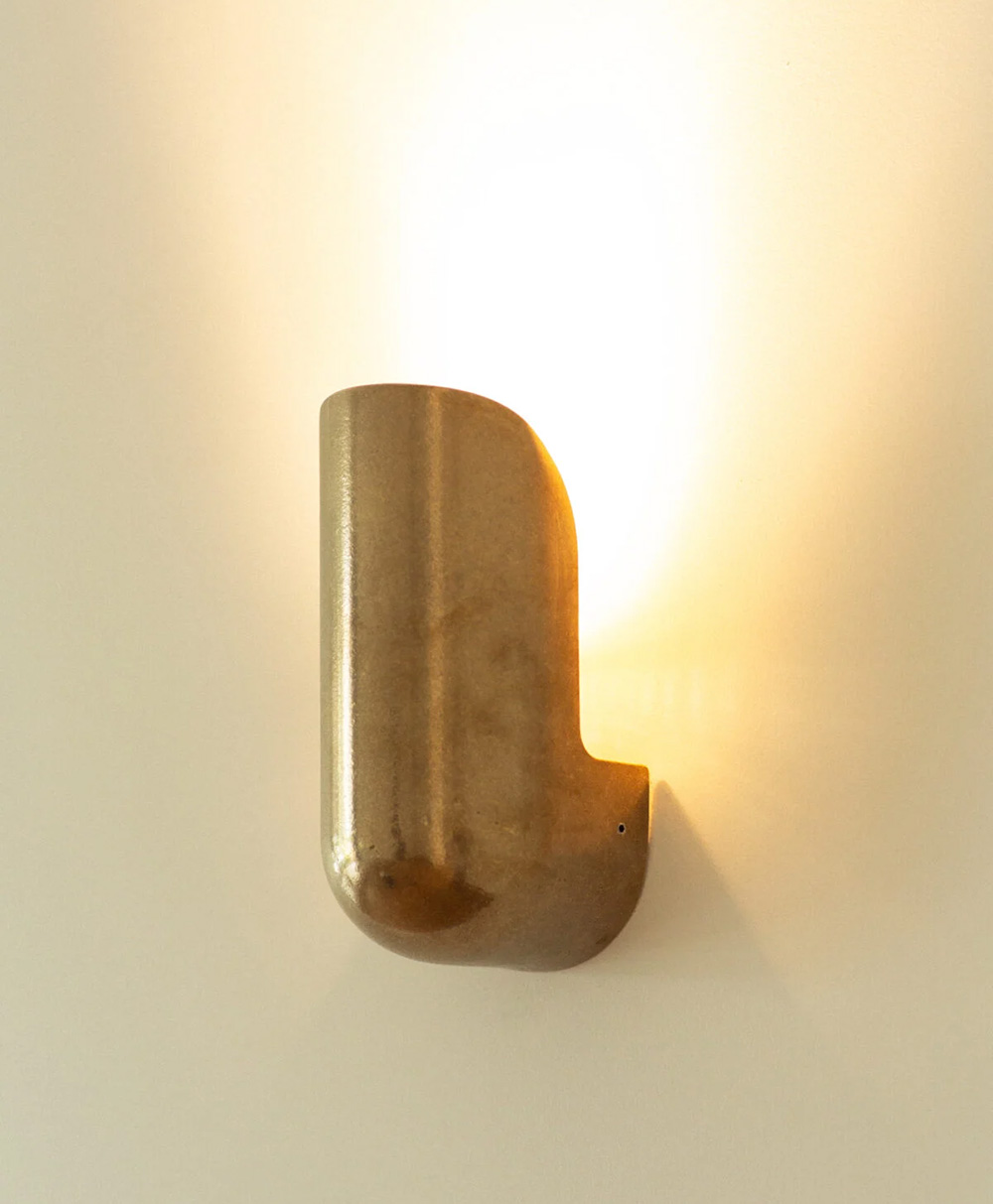 A modern wall sconce with a curved, metallic bronze finish mounted on a white wall, casting a warm, ambient light upward.