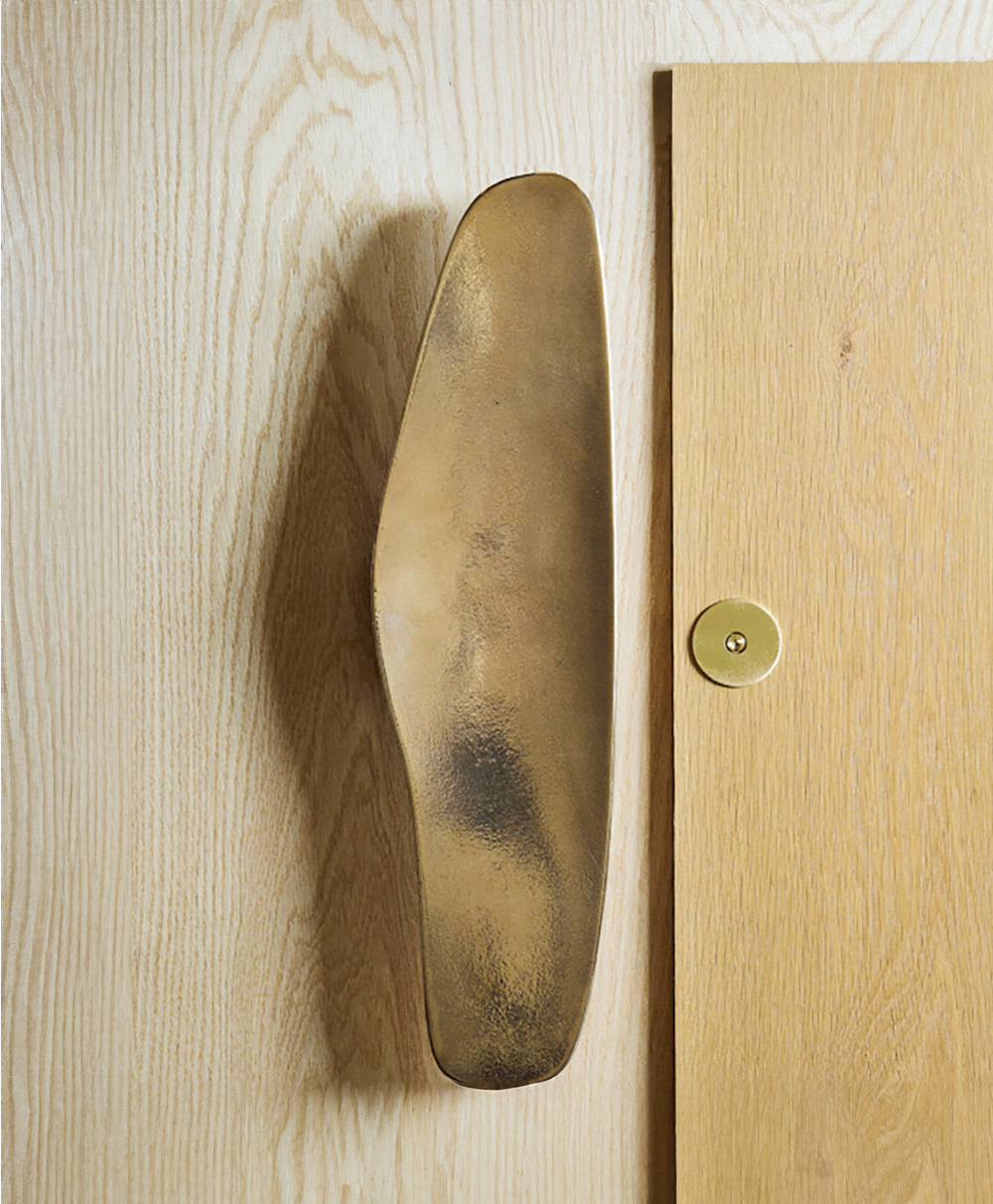 A large, sculpted brass door handle is mounted on a light wood door with a natural grain pattern and a round, brass keyhole plate nearby.