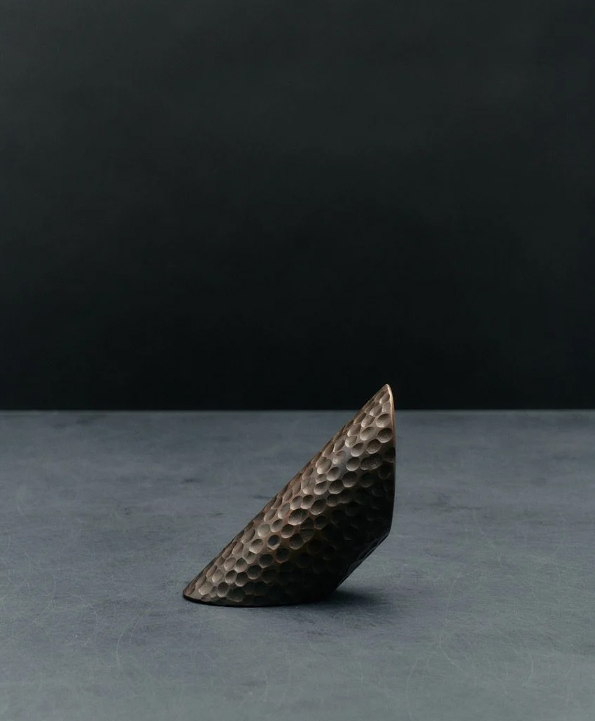 A dark, textured, wedge-shaped object with a hammered surface stands upright on a smooth, gray surface against a dark background.