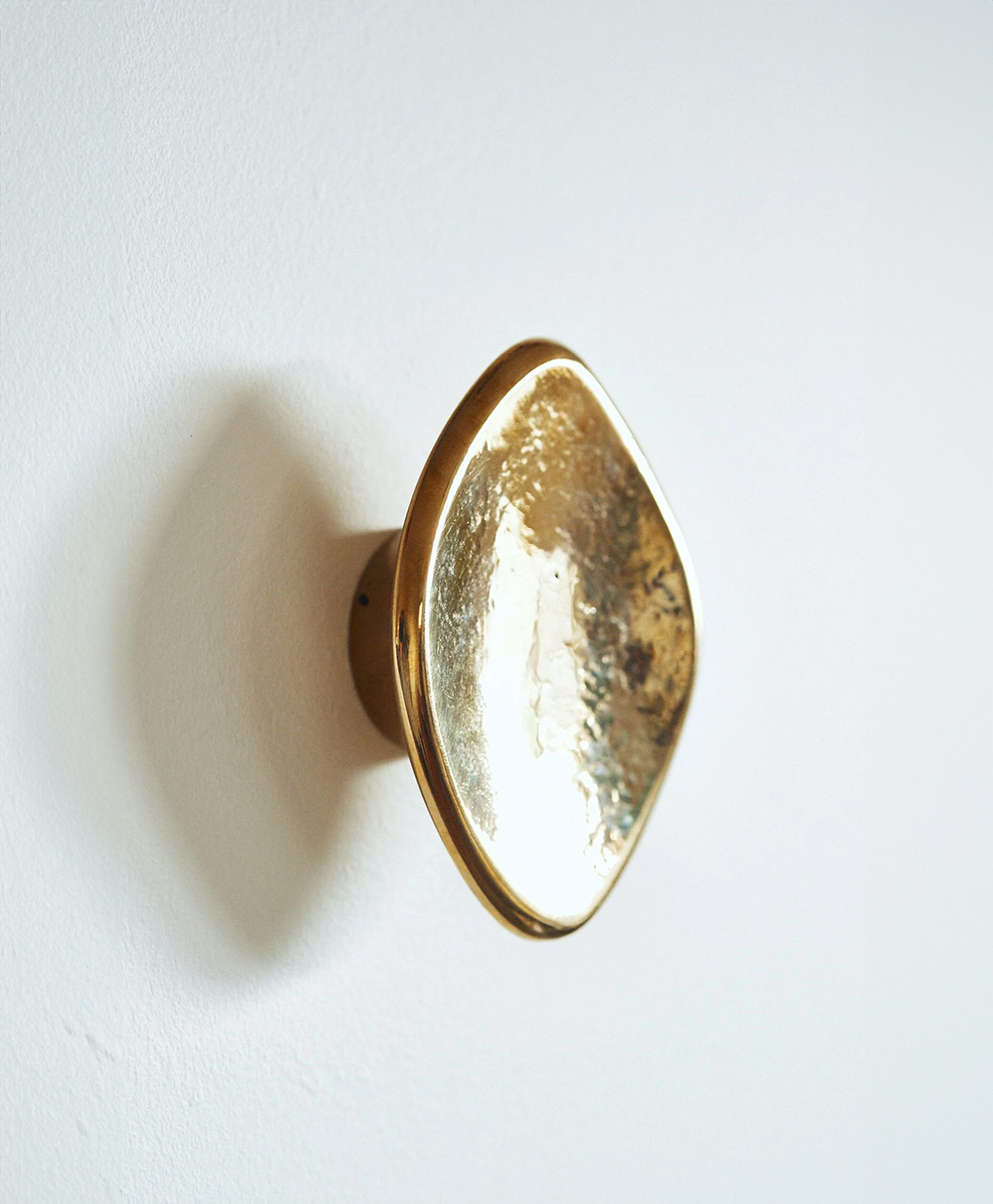 A shiny, gold, leaf-shaped wall hook with a textured surface is mounted on a plain white wall, casting a faint shadow to the left.