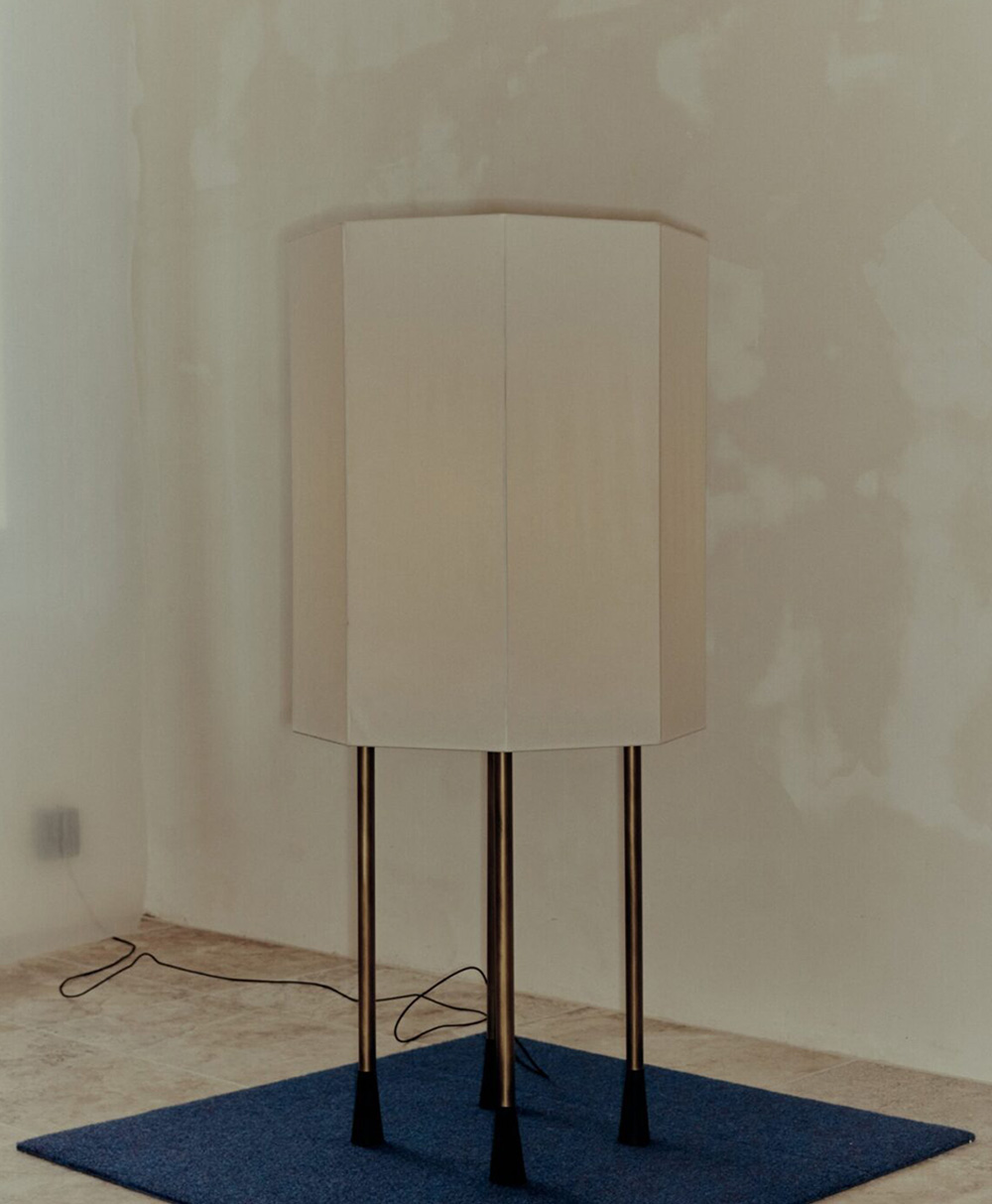 A tall, hexagonal white lampshade rests on four thin black legs, standing on a blue rug in a minimally decorated room with unfinished beige walls and a visible electrical cord on the floor.