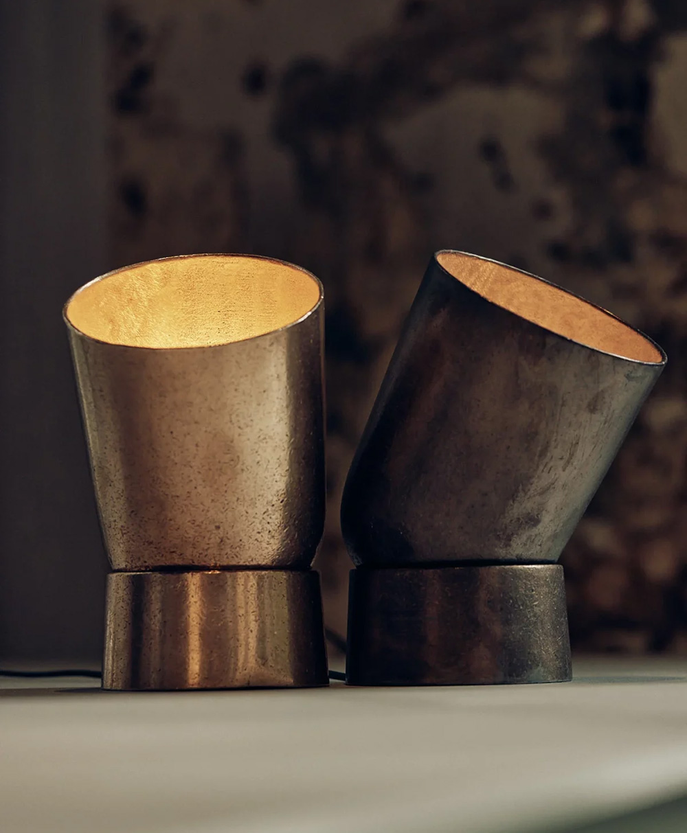 Two modern, metallic table lamps with angled bases and wide, cylindrical shades emit a warm light. The lamps are placed side by side on a surface against a blurred, textured background.