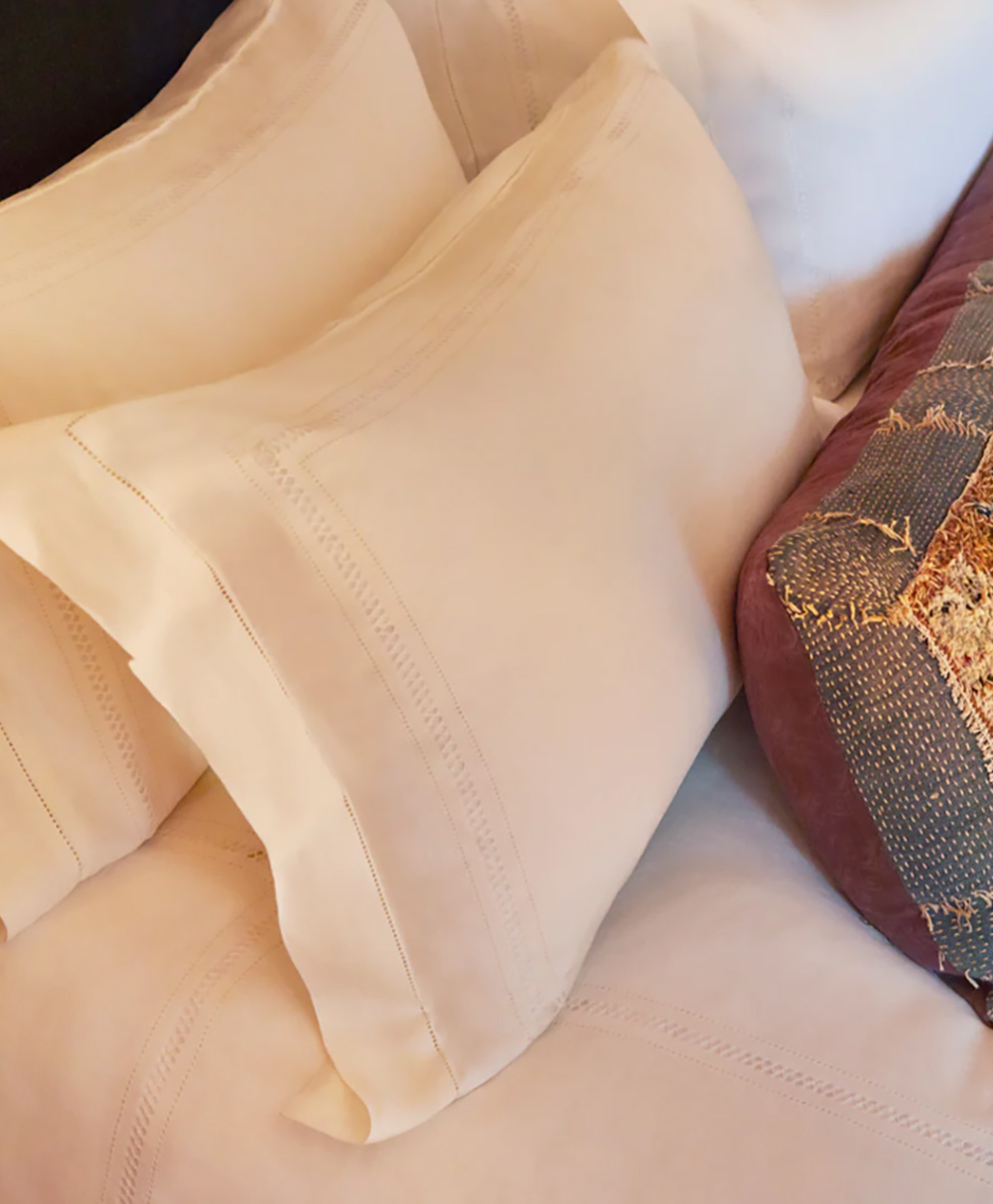 Close-up of a neatly made bed with cream-colored pillows and sheets, featuring detailed stitching. A section of a brown textured blanket or throw is visible on the right side of the image.