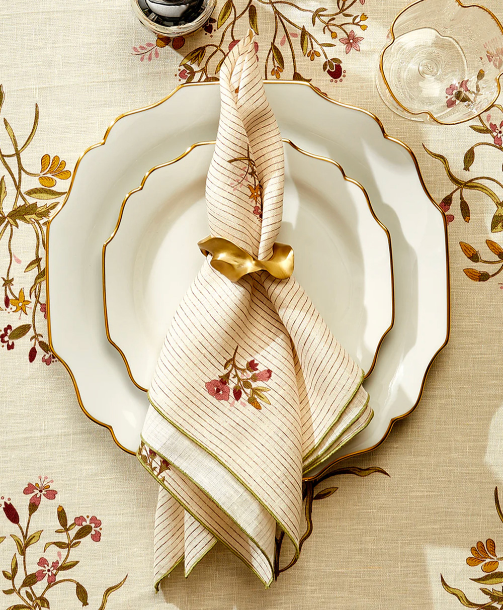 A scalloped-edge white plate is set with a floral-striped napkin tied with a gold ring. The tablecloth has a matching floral pattern, and a wine glass is partially visible in the corner.