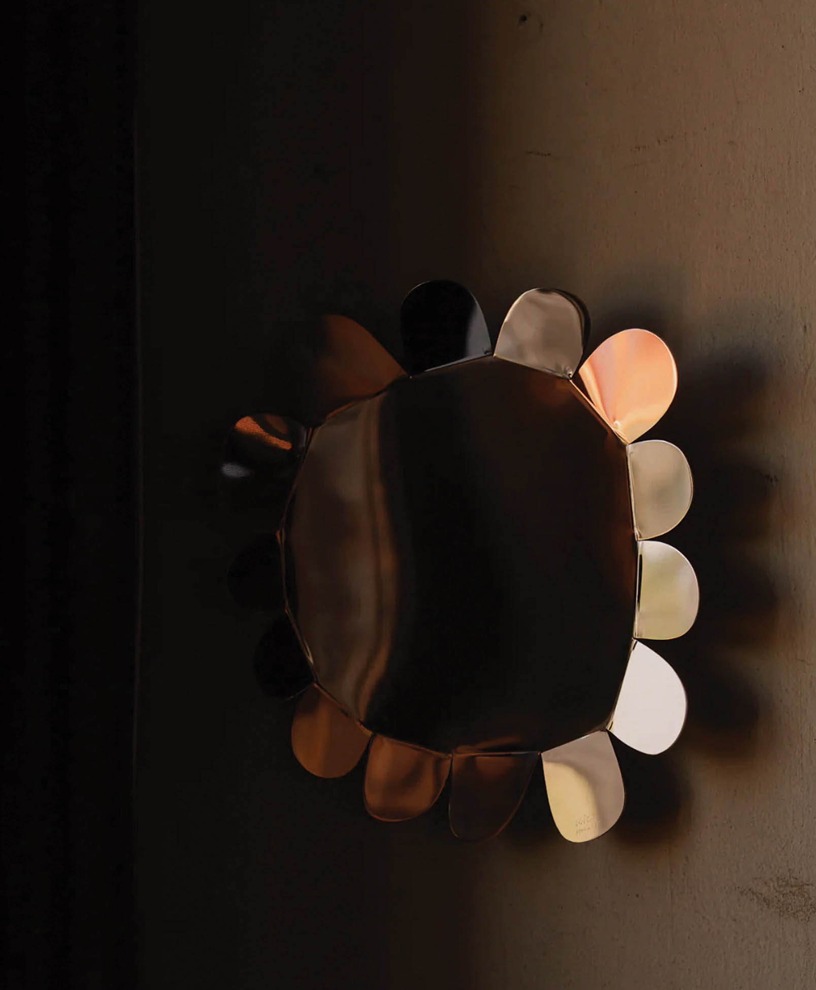 A decorative wall mirror with petal-shaped edges reflecting warm, ambient light, creating soft, colorful glows on a beige wall in a dimly lit room.