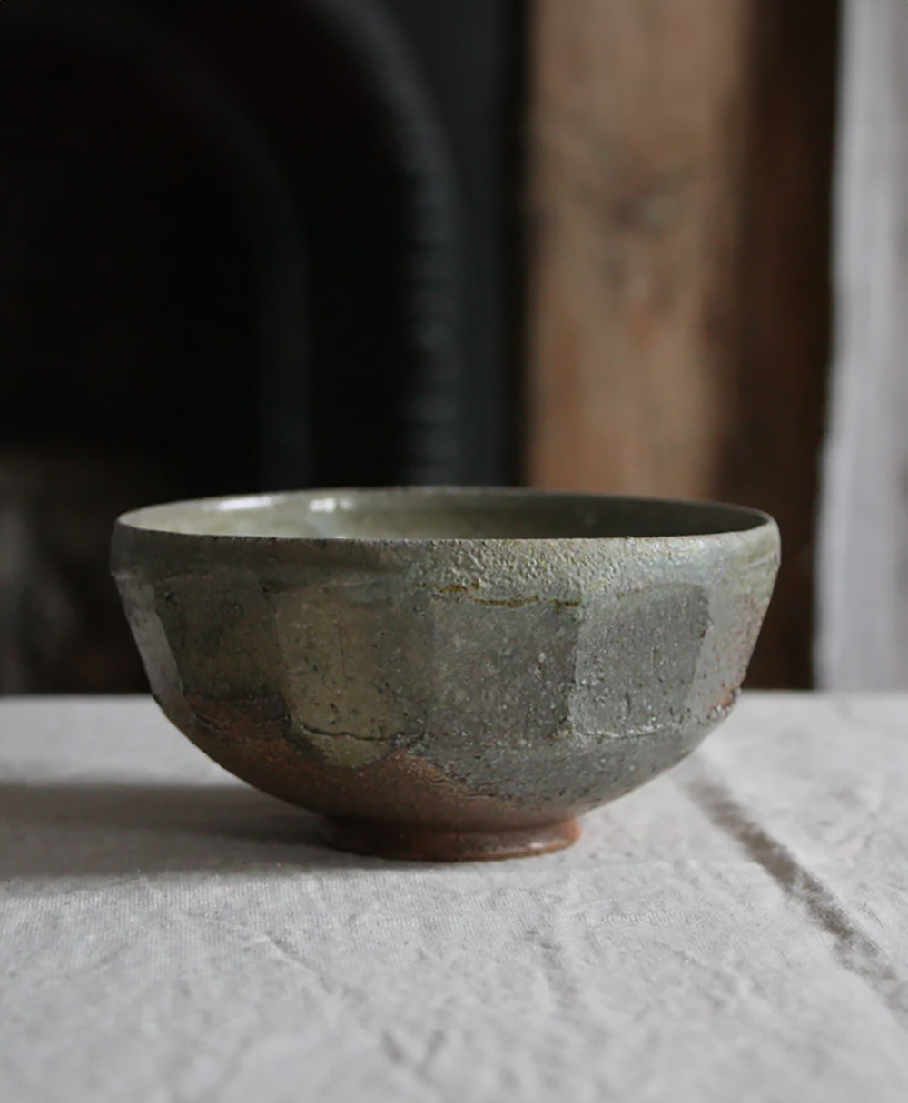 A rustic, handmade ceramic bowl with a textured, earthy glaze sits on a light-colored fabric surface, with a softly blurred background.