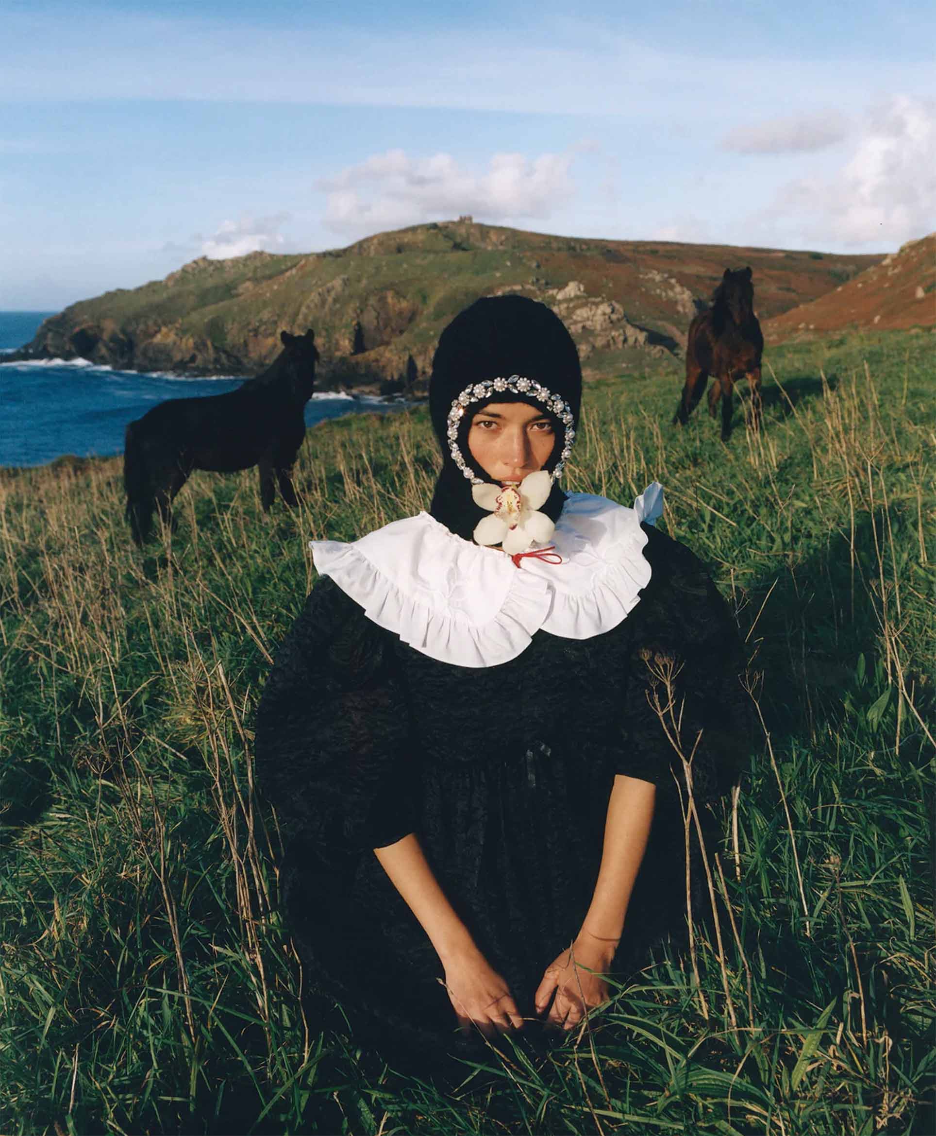 A person in a black dress with a white ruffled collar and a floral headpiece sits in tall grass, holding white flowers in their mouth. Two dark horses stand in the background near rolling hills and a blue sea.