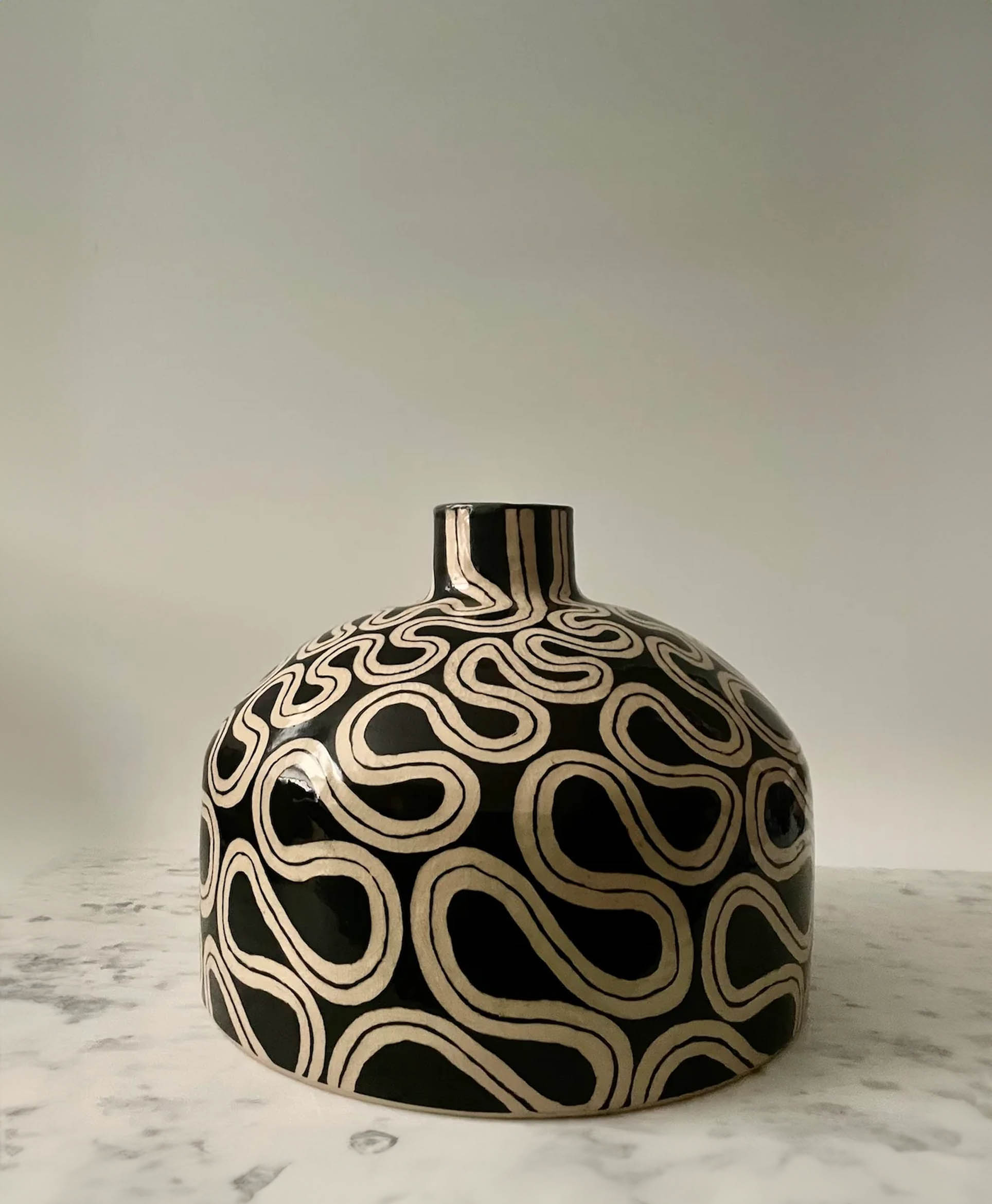 A round, black ceramic vase with a narrow neck sits on a marble surface. The vase features a cream-colored, wavy linear pattern covering its surface, creating a modern, abstract design. The background is plain and light-colored.