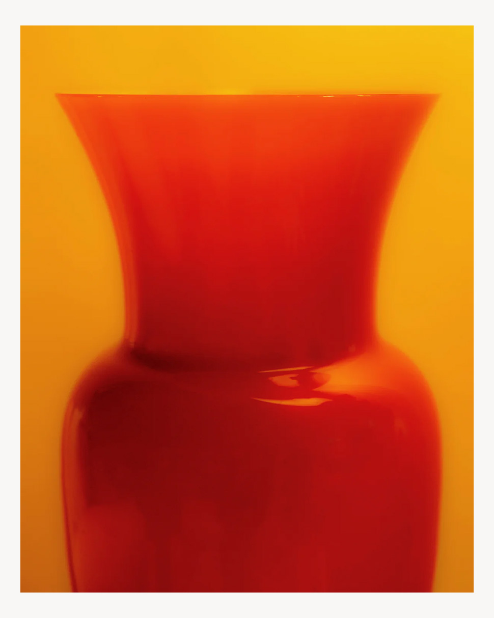 A glossy, bright red vase with a wide rim sits against a vibrant yellow background, creating a bold and colorful contrast.