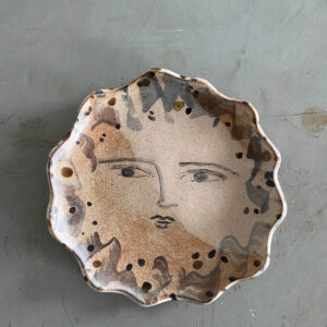A ceramic plate with wavy edges, featuring a painted abstract face with calm eyes and lips, surrounded by brown and black speckled patterns on a light background.