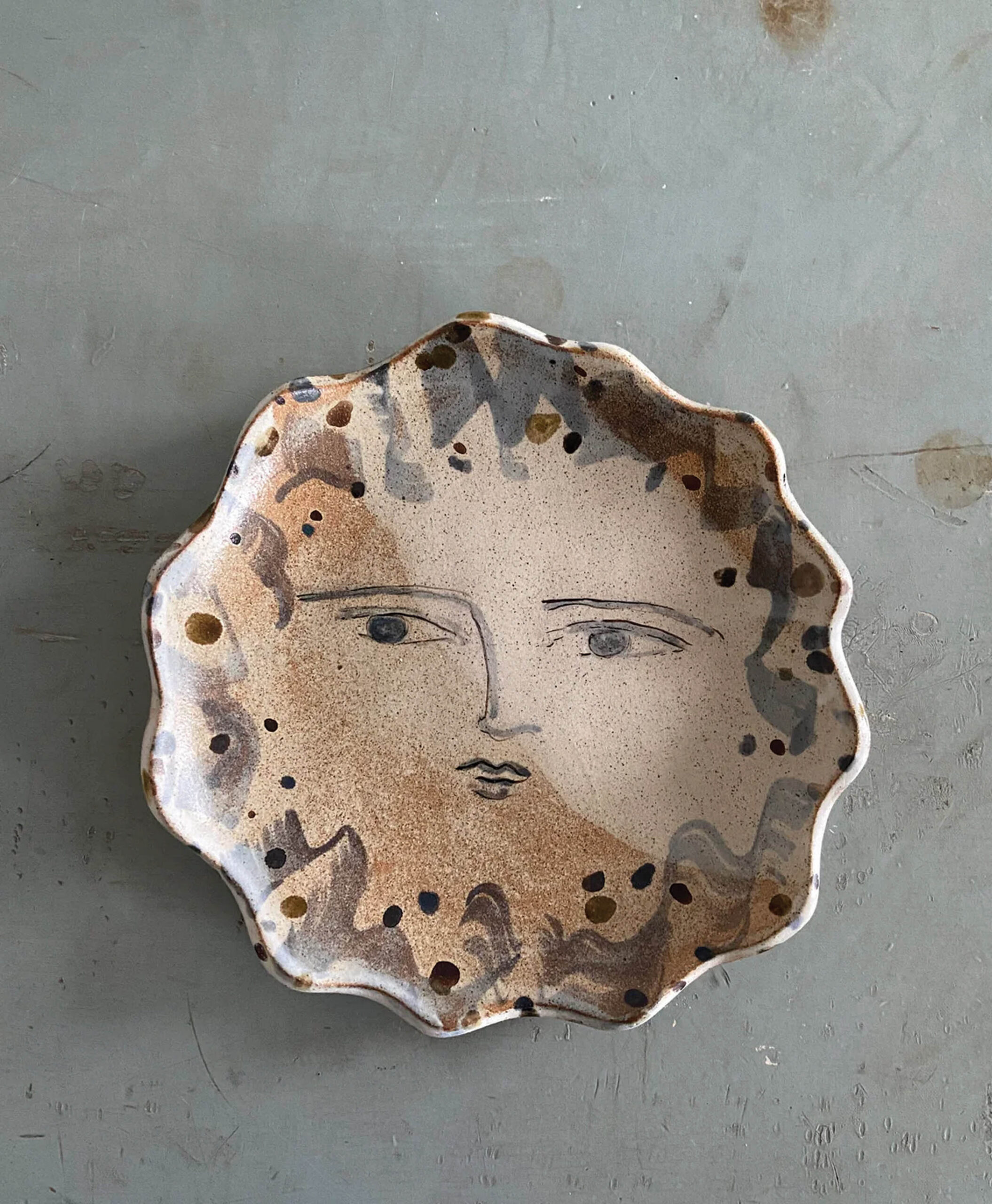 A ceramic plate with wavy edges, featuring a painted abstract face with calm eyes and lips, surrounded by brown and black speckled patterns on a light background.